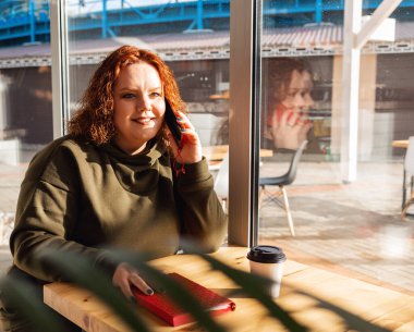 Portrait of a plus dized smiling woman sitting in a cafe and calling at the cellphone. The concept of business and freelancing.