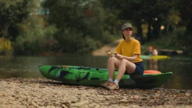 Pretty young woman is sitting on a kayak and have a lunchtime. Real time. A kayak floats in the background. The concept of outdoor activities and picnic.