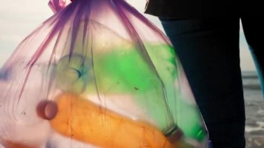 A female volunteer stands on the oceans coast holds a trash garbage bag with plastic bottles. Zoom out. Concept of clean up ocean and ecology.