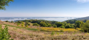 Arka planda uzun adalar bulunan, yüksek eğimli geniş düz nehir kıyısı kısmen sonbaharın başlarında ormanla kaplıdır.