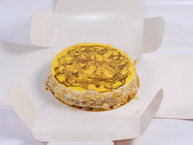 Üzerinde sarı jöle süslemesi olan yuvarlak pasta açık karton ambalajda parçalanmış fındık serpiştirilmiş.