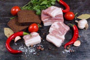 Sebzeler, baharatlar ve yeşillikler arasında yağlı ve yağsız et katmanları ile çiğ domuz eti dilimleri, siyah bir yüzey üzerinde kahverengi ekmek dilimleri