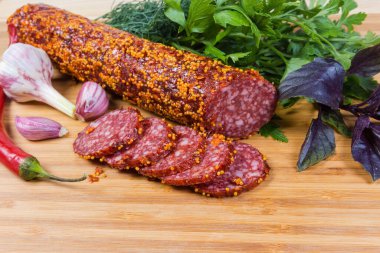 Kısmen dilimlenmiş salam, ahşap kesim tahtasındaki sarımsak, acı biber ve yeşillikler arasında sarı tam tahıllı Fransız hardalı.