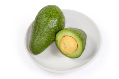 One whole and unpeeled half of the ripe green avocado fruit on a white saucer on a white background