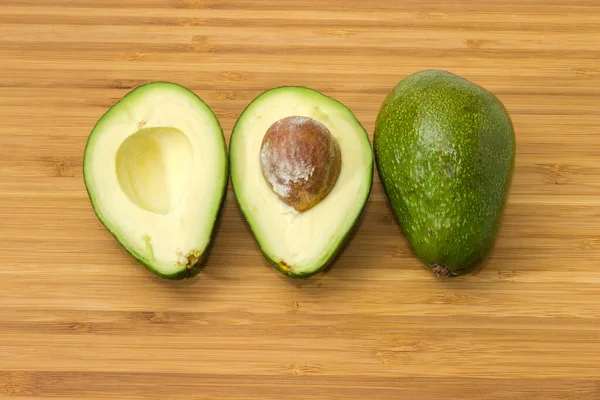One whole and one halved pear-shaped avocado fruits with green skin, top view on a wooden surface