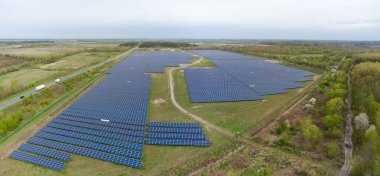 Sabit fotovoltaik panelleri olan yere monte edilmiş güneş enerjisi istasyonu bahar ormanı ve bulutlu havalarda yol, hava panoramik manzarası