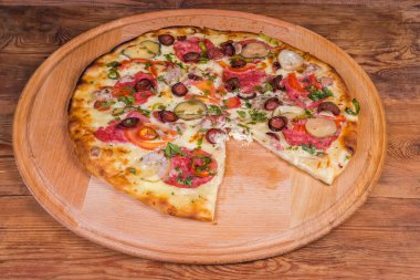 Salam ve diğer sosis ve sebzelerden yapılmış fırınlanmış yuvarlak pizza eski ahşap masadaki büyük yuvarlak ahşap pizza tahtasından bir dilim çıkarıldı.