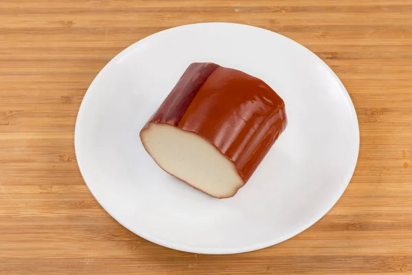 Piece of smoked processed cheese in the form of a cheese sausage part with traditional yellowish-brown outer pellicle on the white dish on a wooden surface