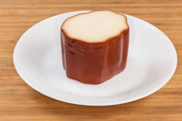 Piece of smoked processed cheese in the form of a cheese sausage part with traditional yellowish-brown outer pellicle on the white dish on a wooden surface