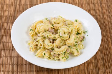 Prepared short curved ruffled pasta, so-called Creste di galli or Rooster comb, mixed with stuffing of boiled chopped pork and fried onion on a white dish on a bamboo table mat