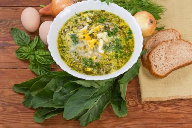 Bowl of the sorrel soup with chopped hard-boiled egg and sour cream among the ingredients for its preparation and slices of the bread on an old rustic table, top view