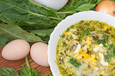 Fragment of bowl of the sorrel soup with chopped hard-boiled egg among the ingredients for its preparation on an old rustic table, top view