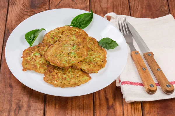 Fried small thick savory zucchini pancakes on the white dish, cutlery on napkin on the old rustic table