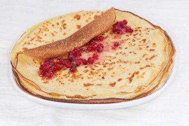 Stack of thin flat round pancakes, the top one of which during roll up into a roll with red currants jam on a dish on a tablecloth, close-up in selective focus