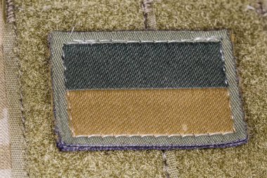 Textile Ukrainian flag sticker in camouflage drab and tan color scheme on an olive base with hook-and-loop fastener fixed on a military camouflage uniform, close-up in selective focus