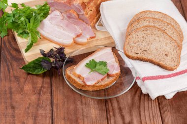 Open sandwich made with grain bread and slice of boiled smoked pork belly on skin, decorated with parsley leaf on the glass saucer against the ingredients on the rustic table