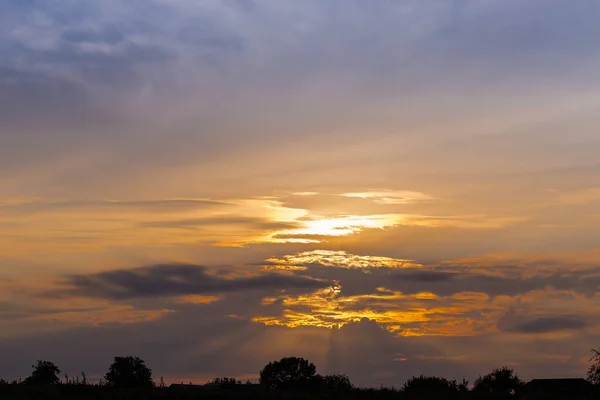 Fragment of the sky with clouds over the silhouettes of trees and roofs of houses on a horizon at sunset