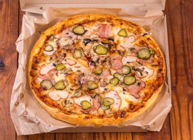 Round pizza with different ham, mushrooms and pickled cucumbers served in open cardboard pizza box on the old rustic table