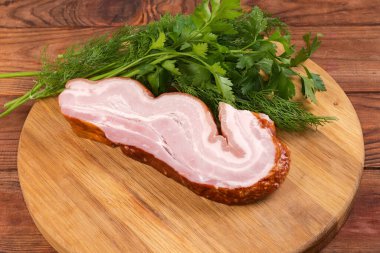 Piece of boiled smoked pork belly and fresh greens on the round cutting board on the rustic table