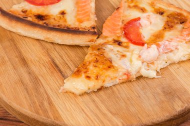 Slices of pizza with salmon and shrimp tails on the pizza serving board on the rustic table, fragment close-up in selective focus