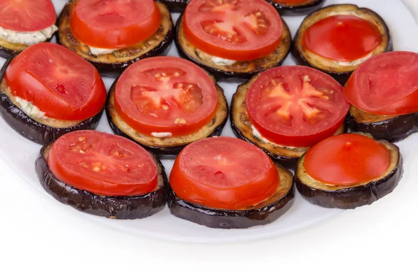 Roasted chopped by circles eggplants with garlic sauce and covered by cut circles of fresh tomatoes on dish, fragment on a white background