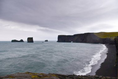 Vast beach with black volcanic sand with coastline on the Atlantic Ocean and an isolated rock in the middle of the sea and a cliff as a backdrop