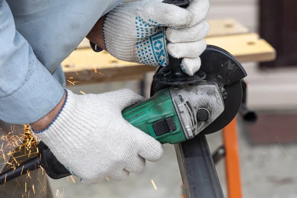 A man in the safety gloves is cutting metal using a green angle grinder tool with orange sparks on a work bench. Horizontal