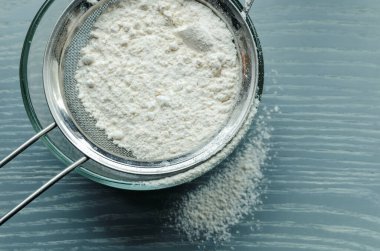 Wheat flour is sown through a sieve into a glass bowl through a metal sieve, on a wooden table.