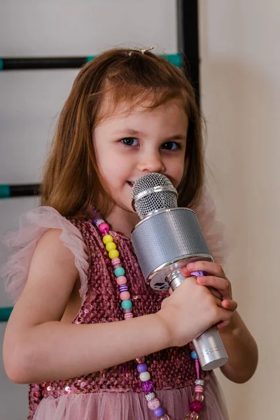 Seorang gadis kecil dalam gaun yang indah bernyanyi ke mikrofon di rumah. Stok Gambar