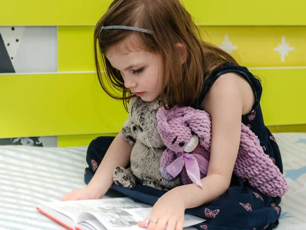 一个小女孩正在床上为她的玩具读一本童话书. 图库图片