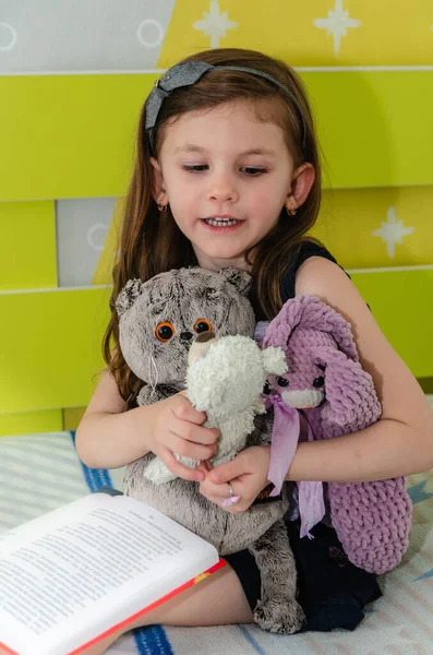 Una niña está leyendo un libro de cuentos de hadas a sus juguetes en su habitación en la cama. Fotos de stock libres de derechos