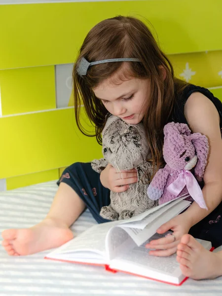 Seorang gadis kecil membaca buku dongeng untuk mainannya di kamarnya di tempat tidur. Stok Foto