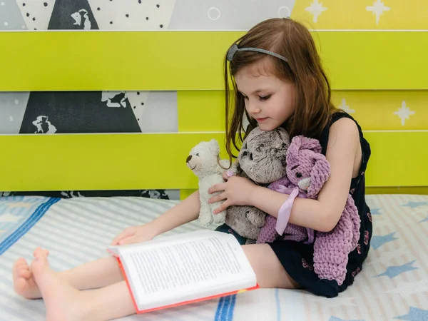 Una niña está leyendo un libro de cuentos de hadas a sus juguetes en su habitación en la cama. Imagen de archivo