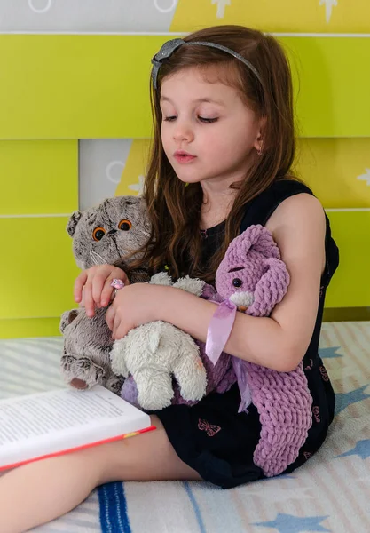 Una niña está leyendo un libro de cuentos de hadas a sus juguetes en su habitación en la cama. Fotos de stock