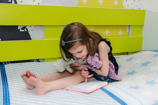 Una niña está leyendo un libro de cuentos de hadas a sus juguetes en su habitación en la cama. Imágenes de stock libres de derechos