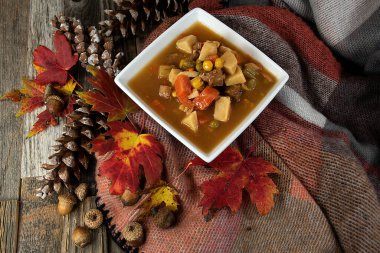 Sonbahar yaprakları, kozalaklar ve meşe palamudu içeren yumuşak bir battaniyede kare beyaz bir kasede sığır eti çorbası.