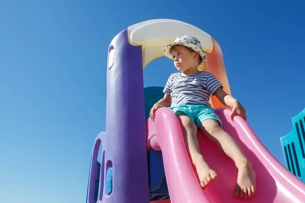 A little boy in a hat is ready to slide, on a colourful slide, he looks into the distance. There is free space for text in the background of the blue sky