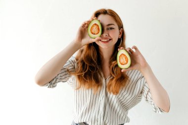 Kızıl dalgalı saçlı, beyaz arka planda kesilmiş bir avokado tutan ve kameraya bakan güzel bir kadının portresi.