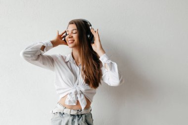 Beyaz gömlekli, kot pantolonlu, beyaz arka planda poz veren güzel esmer kadın. Genç yaşam tarzı konsepti. Kulaklıkla müzik dinlemek.