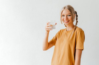 Sarı tişörtlü sıradan bir kadın beyaz stüdyo arka planında poz veriyor. Elinde su bardağı var.