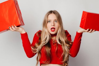 Kırmızılı genç sarışın kadın elinde hediyelerle kameraya şüpheli ve şüpheci bir ifadeyle bakıyor.