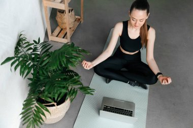 Çevrimiçi eğitim kavramı. Genç, sıska kadın internette yoga yapıyor. Paspasın üzerinde oturuyor. Önünde dizüstü bilgisayar var.
