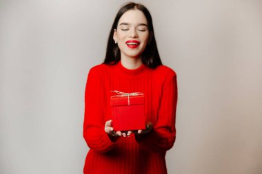 Şirin kız hediyesini göstererek gülümsüyor. Kutu orta büyüklükte, kırmızı ve kurdeleli.