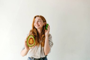 Kızıl dalgalı saçlı, beyaz arka planda kesilmiş bir avokado tutan ve kameraya bakan güzel bir kadının portresi.