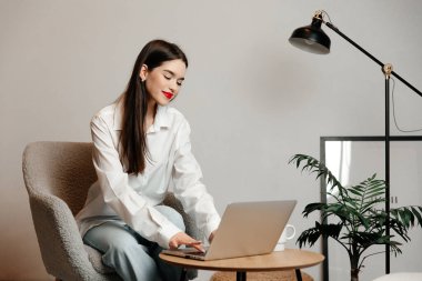 Çekici genç bir kadın arka planda rahat bir sandalyede oturuyor. Kızın siyah saçları, kırmızı dudakları ve beyaz gömleği var.