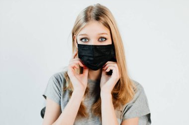 Mavi gözlü kız kendini hastalıktan korumak için yüz maskesi takıyor. Genç bir kadın arka planda tek başına duruyor.