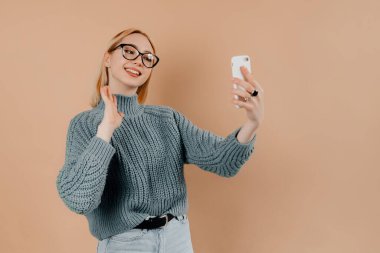 Şirin kız blogu için selfie çekiyor. Kız tarafsız renklerden ve büyük gözlüklerden oluşan günlük giysiler giyiyor.