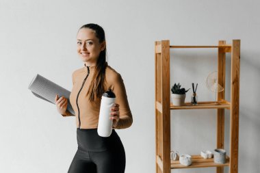 Spor elbiseli, at kuyruklu, yoga minderi ve su şişesi tutan gülümseyen kadın gününü düşünüyor.