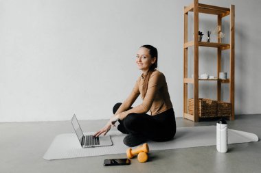 Sağlıklı bir kadın paspasın üzerinde oturuyor, egzersiz yapmaya hazır, dizüstü bilgisayara bakıyor. Sağlıklı yaşam tarzı ve kendini motive etme kavramı