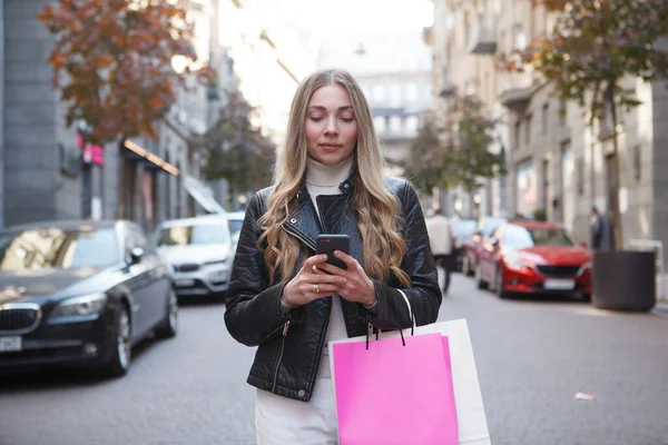 美しいですスタイリッシュな女性とともにショッピングバッグテキストメッセージ上の彼女のスマートフォン立って路上 — ストック写真